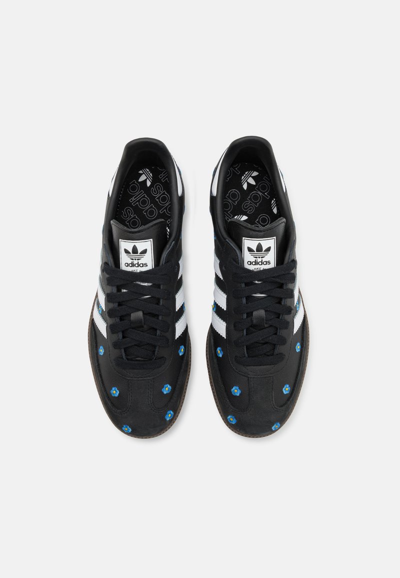 Zapatillas de ante negro con diseño de tres rayas blancas, acentos florales azules y suela de goma texturizada. Logo en la lengüeta y plantilla con patrón.