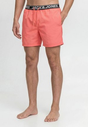Uomo con pantaloncini da bagno color corallo con fascia in vita Jack & Jones nera e bianca, in piedi a piedi nudi con una mano in tasca.