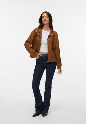 Kvinde iført en brun jakke over en hvid crop top, mørkeblå flared jeans og brune spidse sko, stående mod en hvid baggrund.