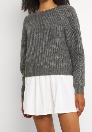 Kvinde iført en grå strikket trøje med lange ærmer og et kort hvidt skørt, vist fra skuldrene til midt på låret.