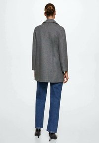 Mujer con el cabello recogido en un moño, vistiendo un abrigo gris, jeans azules y botas de tacón negras, de pie mirando hacia atrás contra un fondo sencillo.