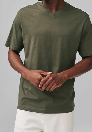Homme portant un T-shirt à col en V manches courtes couleur vert olive, les mains délicatement jointes devant lui, sur un fond gris uni.