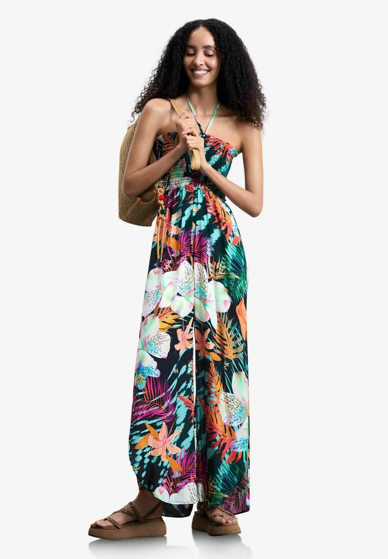 Mujer con cabello rizado que lleva un vestido largo floral colorido, sosteniendo un bolso de hombro tejido y sonriendo con los ojos cerrados.