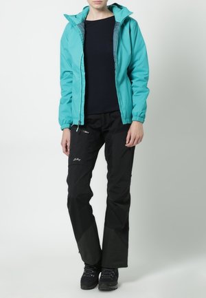 Personne portant une veste à capuche turquoise, un chemisier noir à manches longues, un pantalon de plein air noir avec une poche zippée et des chaussures de randonnée noires.