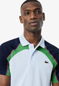 Polo con corpo azzurro chiaro, maniche blu navy e un pannello testurizzato verde con rifiniture rosa. Include un piccolo logo verde sul petto.