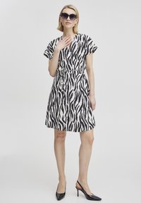 Zebra-muster Kleid in Schwarz und Weiß, mit kurzen Ärmeln, tailliertem Schnitt und ausgestelltem Rock. Getragen mit schwarzen High-Heels.