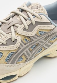 Sneakers med en kombination av mjuk beige mocka och mesh, med grå accenter och ljusblå detaljer, som visar en andningsbar och strukturerad design.