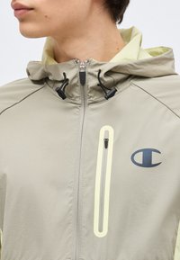 Άτομο που φοράει μπεζ φερμουάρ με κουκούλα και μαύρα κορδόνια, με κάθετη τσέπη με φερμουάρ και λογότυπο Champion στο στήθος.