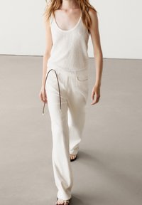 Un débardeur blanc à côtes associé à un pantalon large texturé blanc avec un cordon de serrage marron. Des sandales complètent la tenue.