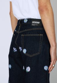 Jeans de mezclilla oscura con intrincados bordados florales en azul y rosa. Incluye una etiqueta de marca en la cintura y un diseño clásico de bolsillos.