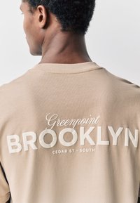 Béžové bavlnené tričko s grafickým motívom na zadnej strane s nápisom "Greenpoint" kurzívou a "BROOKLYN" tučnými 3D písmenami. Hladká textúra, okrúhly výstrih.