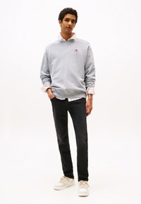 Svetlo siva jopa z okrolim ovratnikom in majhnim logotipom, nošena čez roza in belo črtasto majico. Temne jeans hlače in svetli športni copati dopolnjujejo videz.