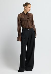 Braune transparente Bluse mit einem Schleifenkragen, kombiniert mit hochtaillierten schwarzen weiten Hosen, die einen dekorativen Taillenknopf haben.