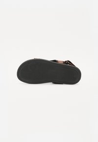 Melnā gumijas zole ar tekstūrētu rakstu; redzama brūna ādas siksna, kas demonstrē plakanu un ergonomisku dizainu.