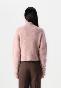 Suéter corto rosa hecho de un material suave y esponjoso; cuenta con un cuello alto, puños y dobladillo acanalados; combinado con pantalones marrones ajustados.