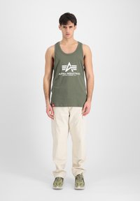 Top cașmir verde măsliniu cu logo-ul alb "ALPHA INDUSTRIES", asortat cu pantaloni cargo bej. Modelul stă cu mâinile pe lângă corp într-o poziție neutră.