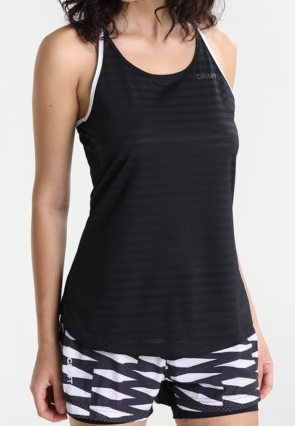 Mujer con camiseta deportiva sin mangas negra y pantalones cortos deportivos con estampado en blanco y negro, de pie con los brazos relajados a los lados.