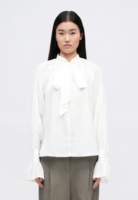 Blusa bianca a maniche lunghe con colletto a fiocco, con polsini delle maniche arricciati e taglio dritto. Tessuto liscio senza motivi o texture visibili.