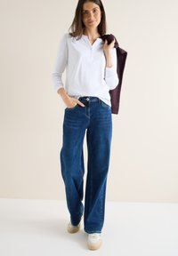 Letthvid langærmet henley-shirt med knapdetalje,/parret med brede mørkeblå jeans og hvide sneakers med beige accenter.