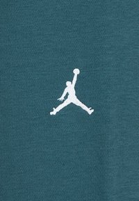 Tamsiai mėlynas audinys su lygiu paviršiumi puošia baltas siuvinėtas Jumpman logotipas, vaizduojantis figūrą, šokantį laikydamas krepšinio kamuolį.
