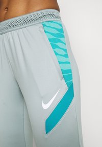 Helle graue Sporthosen mit einem atmungsaktiven Bund, ausgestattet mit einem türkisfarbenen seitlichen Einsatz mit einem wellenförmigen Muster und einem kleinen weißen Logo.