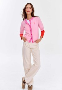 Różowy kardigan z czerwonymi akcentami, zapięcie na guziki i mały detal w kształcie serca. Połączony z jasnobeżowymi spodniami i wzorzystymi butami.