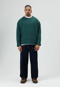 Groen oversized sweatshirt met een ronde hals, gedragen over een wit T-shirt, gecombineerd met donkere spijkerbroek en bruine wandelschoenen.