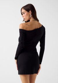 Schwarzes schulterfreies Kleid mit langen Ärmeln, figurbetonter Silhouette und Reißverschluss am Rücken. Glatte Textur und mittellange Länge bis zur Mitte des Oberschenkels.