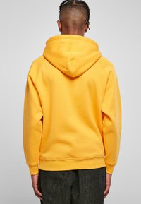 Sudadera amarilla con una gran capucha y puños acanalados, con una textura suave. La vista muestra la parte trasera sin logotipos ni gráficos visibles.