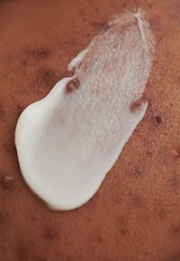 Creamy white lotion is spread on textured brown skin, revealing small bumps and a smooth area where the lotion is applied.