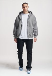 Grauer Kapuzenpullover mit Reißverschluss, großer Kapuze und Vordertaschen, getragen über einem weißen T-Shirt und kombiniert mit schwarzen Hosen und blauen Turnschuhen.