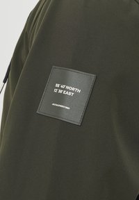 Veste vert foncé avec un patch carré noir affichant les coordonnées 55°40' Nord 12°36' Est et le nom de la marque Jack & Jones.