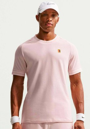 Moški v svetlo roza športni majici, beli Nike kapici in belih zapestnicah stoji pred enobarvnim svetlim ozadjem.