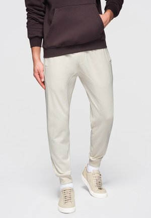 Persona con pantaloni da jogging beige, sneakers beige con suola bianca, calzini bianchi e felpa con cappuccio marrone scuro, in piedi con una mano in tasca.