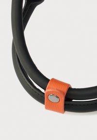 Μαύρες ελαστικές θηλιές με πορτοκαλί λουρί από δέρμα, στερεωμένο με ασημένιο κουμπί. Ομαλή υφή και μινιμαλιστικός σχεδιασμός.