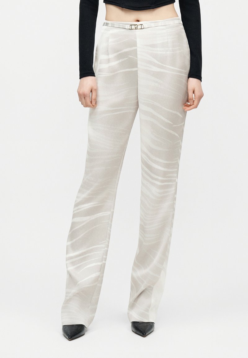 Kvinde iført beige bukser med høj talje og vide ben med diskret hvidt bølgemønster, kombineret med sorte spidse sko og en sort crop-top med lange ærmer.