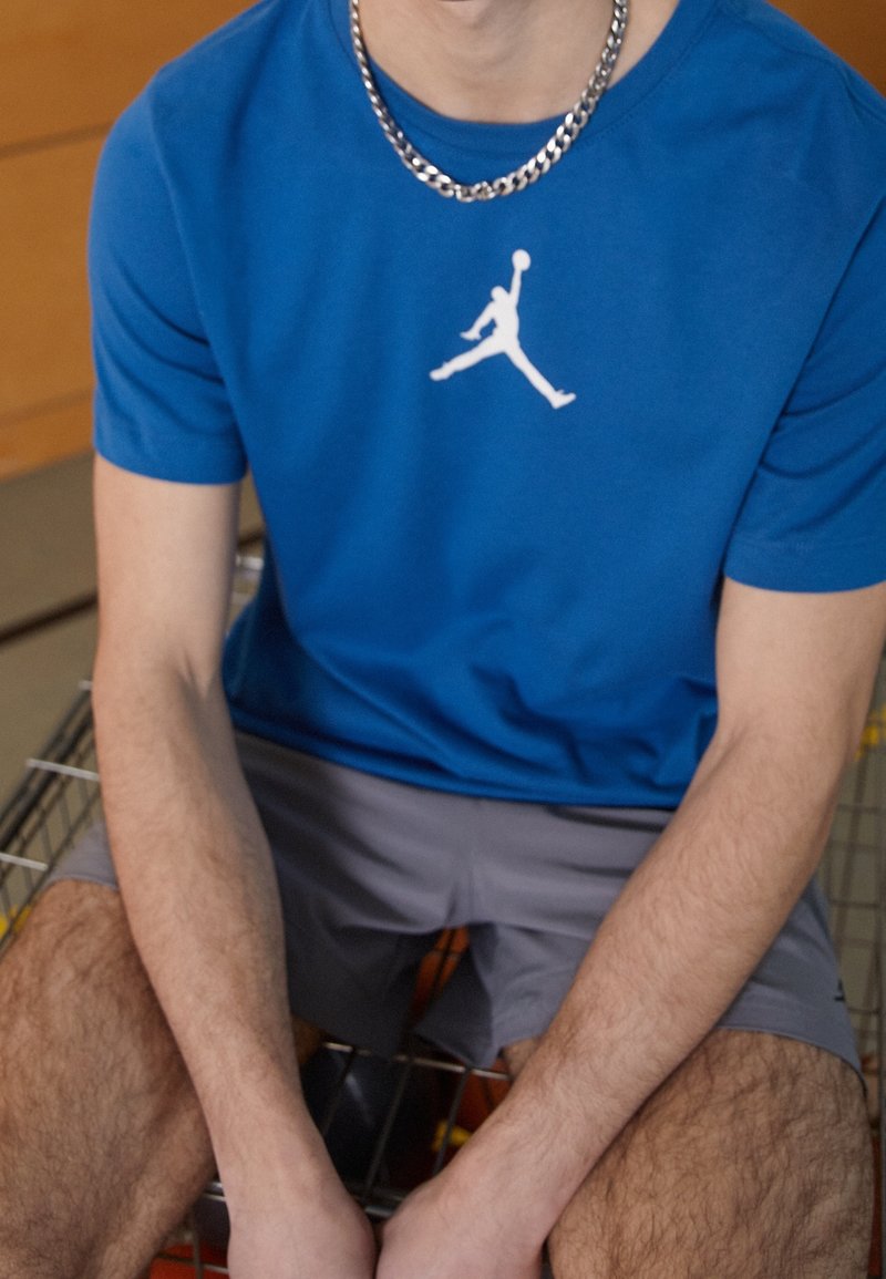 Blå idrottströja med vit Air Jordan-logotyp, tillsammans med grå shorts. Enkelt design, korta ärmar, och ett kedjehalsband synligt.