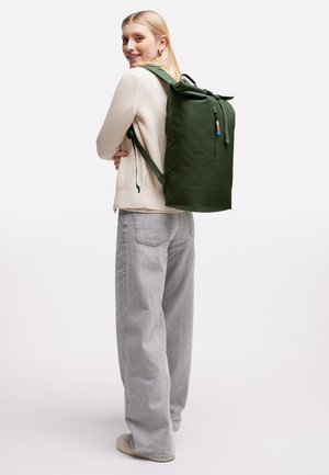 Ung kvinde iført cremefarvet sweater og grå bukser bærer en stor mørkegrøn roll-top rygsæk og smiler over sin skulder.