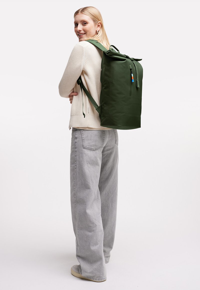 Jeune femme portant un pull crème et un pantalon gris, portant un grand sac à dos roulé de couleur vert foncé, souriant par-dessus son épaule.