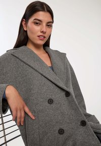 Młoda kobieta z długimi ciemnymi włosami, ubrana w szary podwójnie zapinany wełniany płaszcz, siedząca na metalowym krześle na jednolitym tle.