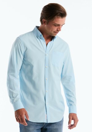 Man draagt lichtblauw overhemd met lange mouwen en borstzak, kijkt naar beneden, tegen een effen witte achtergrond.