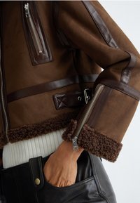 Giacca marrone con accentuazioni in pelle, tasche con cerniera, orlo peloso e materiali testurizzati. Abbinata a un top bianco a coste e pantaloni neri.