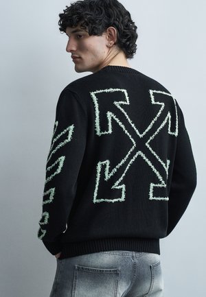 Mladý muž s kučeravými vlasmi, oblečený v čiernom svetri s veľkými bielymi šípkami na chrbte a pozdĺž rukáva, doplnený o vyblednuté džínsy.