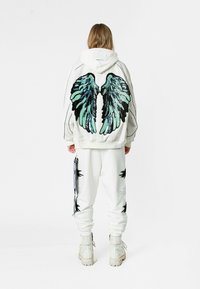 Sudadera blanca con un gran diseño de alas negras y verde azulado en la parte posterior, combinada con pantalones a juego que presentan acentos negros. Botas blancas gruesas completan el look.