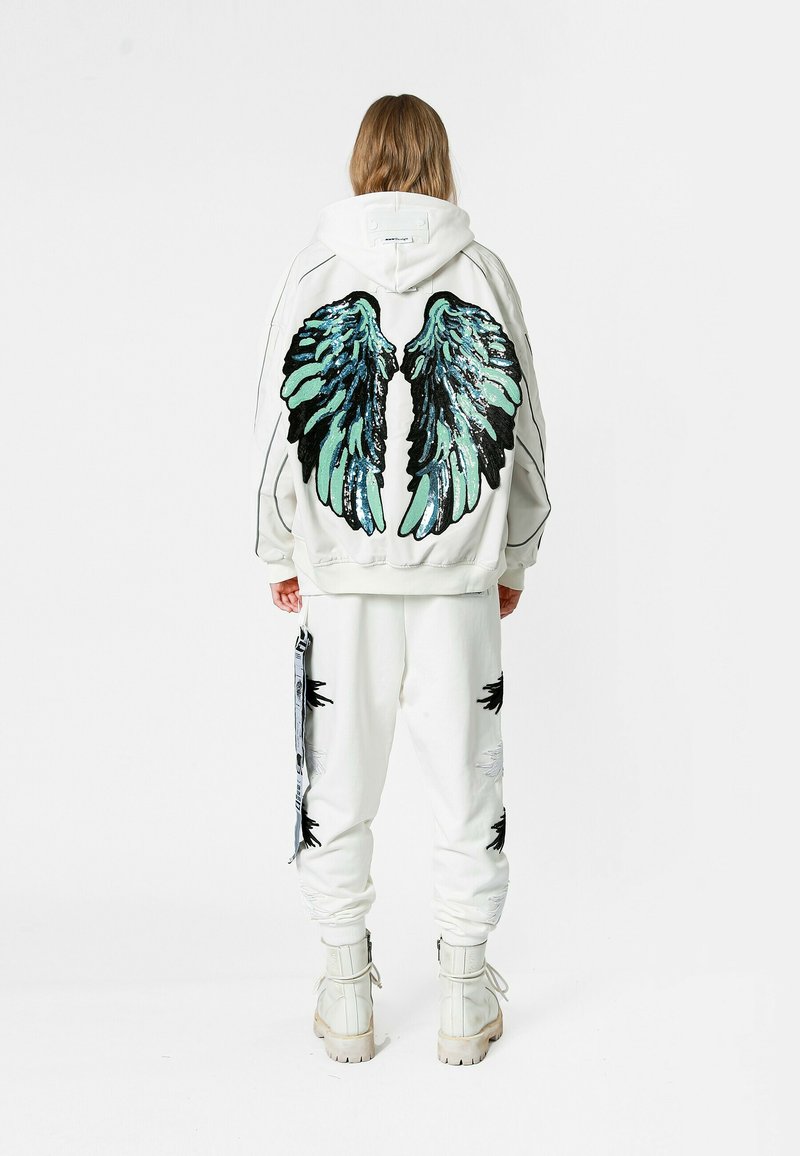 Sudadera blanca con un gran diseño de alas negras y verde azulado en la parte posterior, combinada con pantalones a juego que presentan acentos negros. Botas blancas gruesas completan el look.