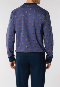 Uomo che indossa una camicia a maniche lunghe blu con un motivo di piccoli camion rossi che trasportano piante verdi, abbinata a pantaloni blu scuro.