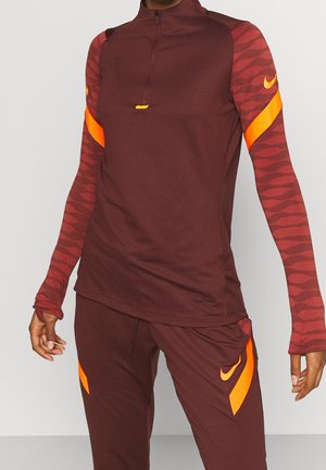 Sportoutfit met een bordeauxrode longsleeve en broek met oranje geometrische patronen en Nike-logo's op de mouwen en dijen.