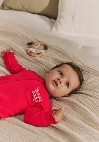 Rød ribbet jumpsuit med hvid "santa baby" tekst, liggende på et beige tekstureret tæppe med en blød hundelegetøj ved siden af. Babyen kigger opad.
