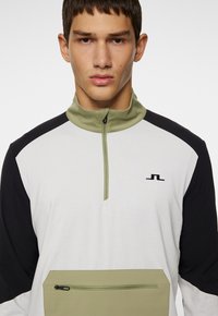 Langærmet pullover i hvid, sort og olivengrøn. Har en lynlås krave, en lomme foran og et logo på brystet.
