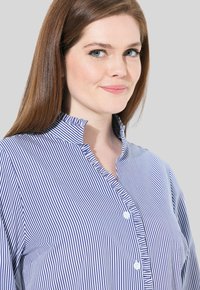 Sininen ja valkoinen raidallinen nappikauluspaita, jossa on röyhelökaulus ja nappilista, ja joka on valmistettu sileästä kankaasta ja se on muotoiltu.